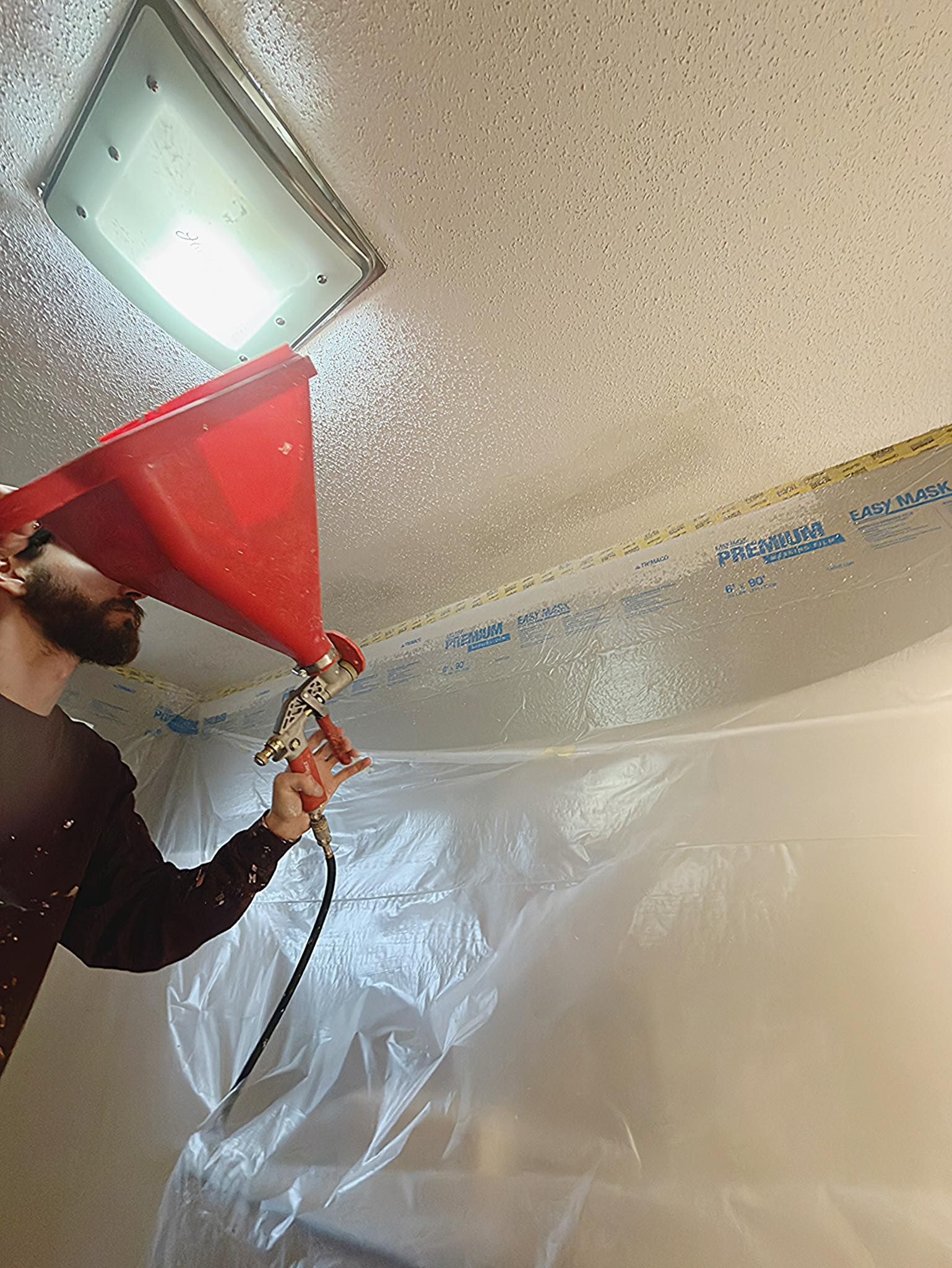 Man spraying textured ceiling with a red hopper and spray gun, plastic sheeting covers a wall.