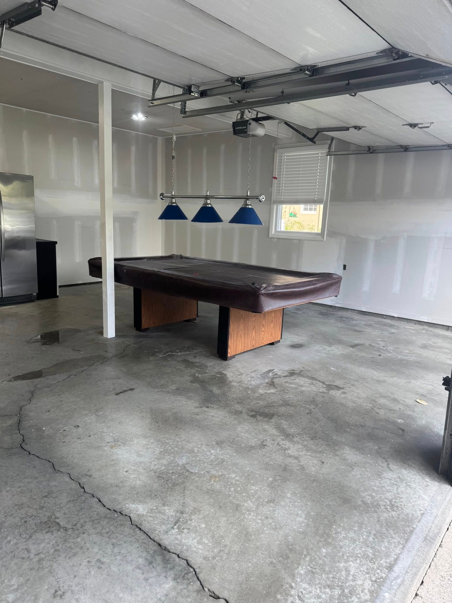 Pool table in a garage, under blue pendant lights. Unfinished walls, concrete floor.