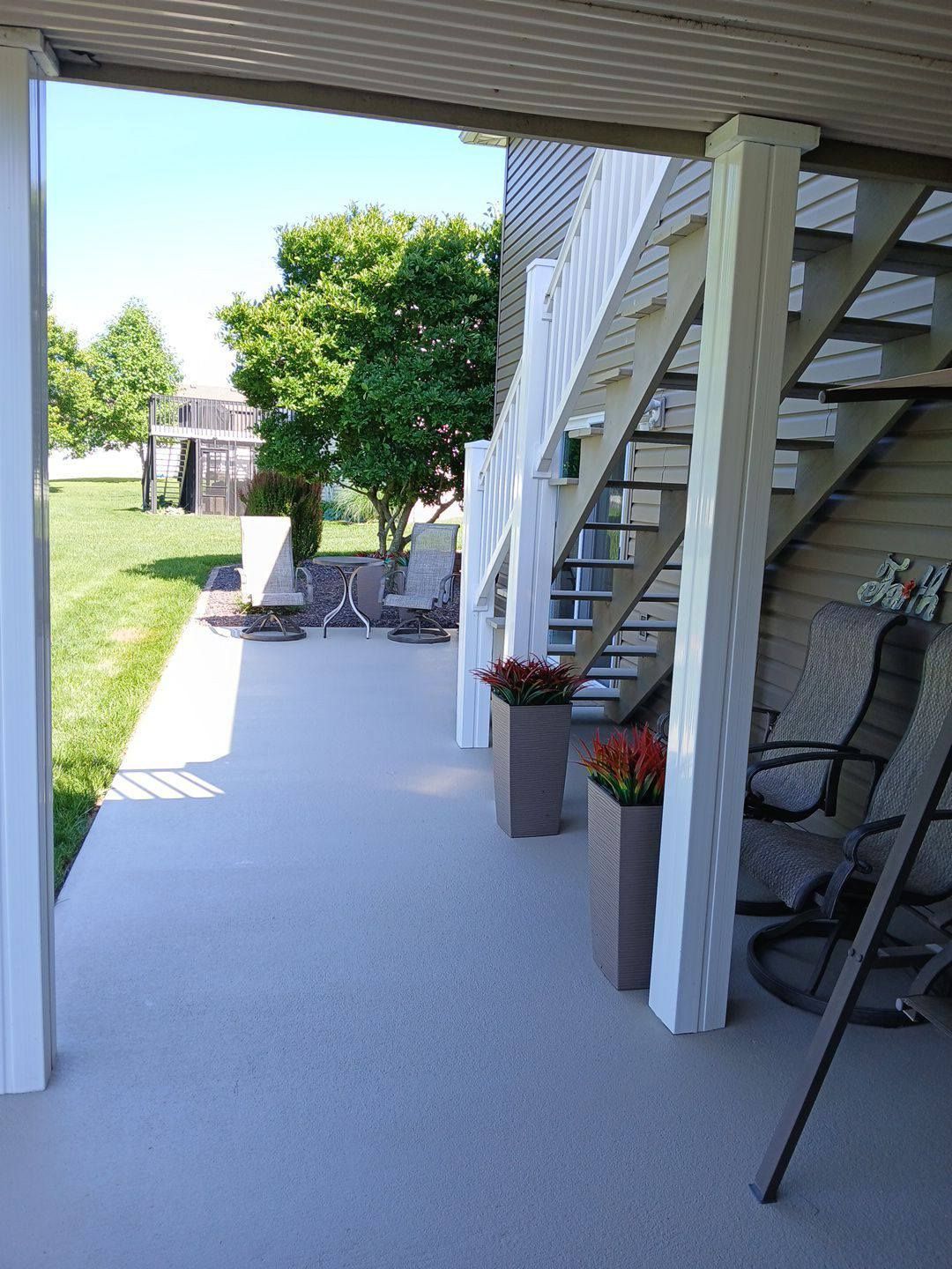 Covered patio with stairs, seating, and potted flowers. Green lawn and trees in the background.