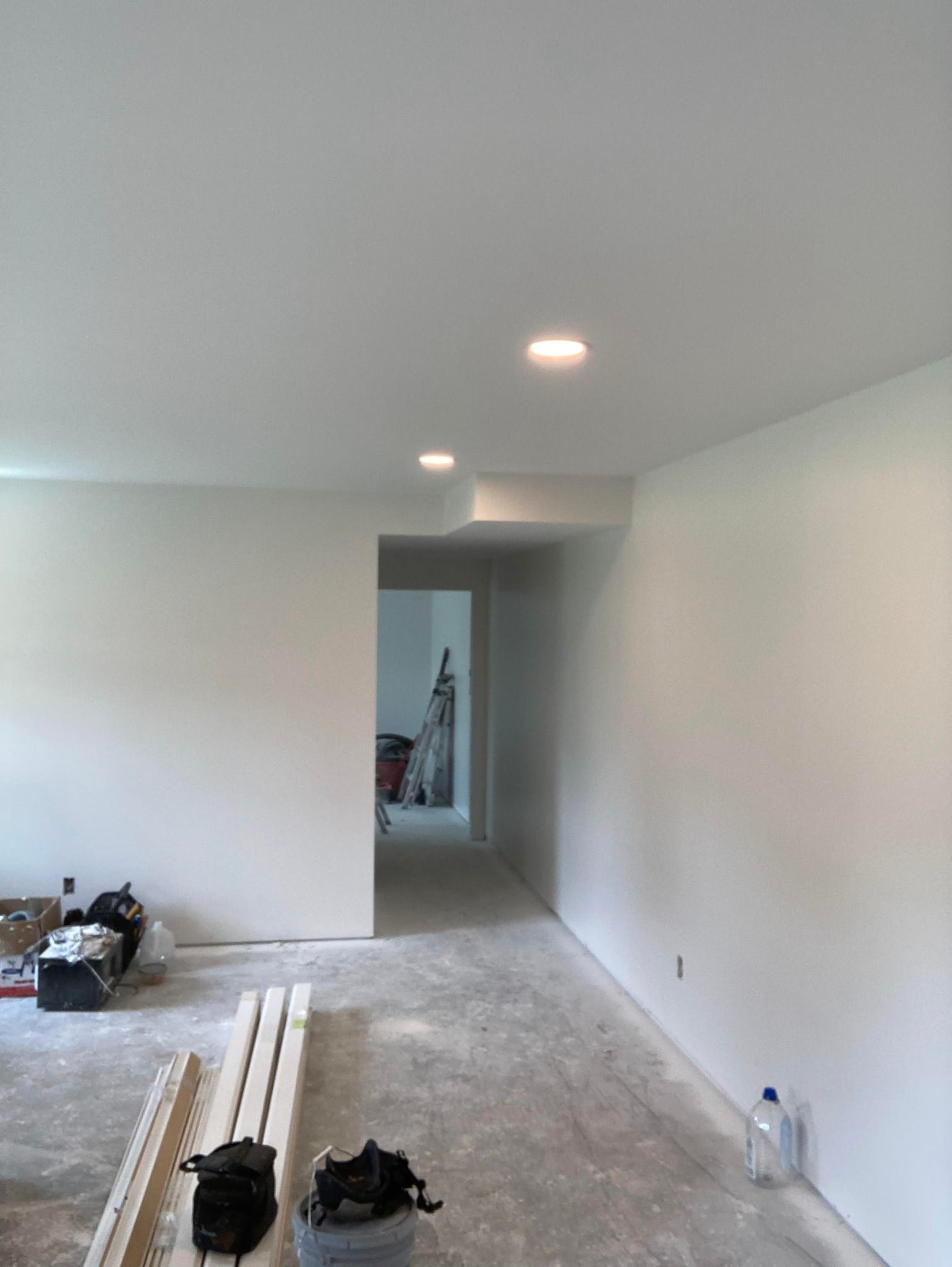 Interior view of a room under construction with white walls, recessed ceiling lights, and an unfinished floor.