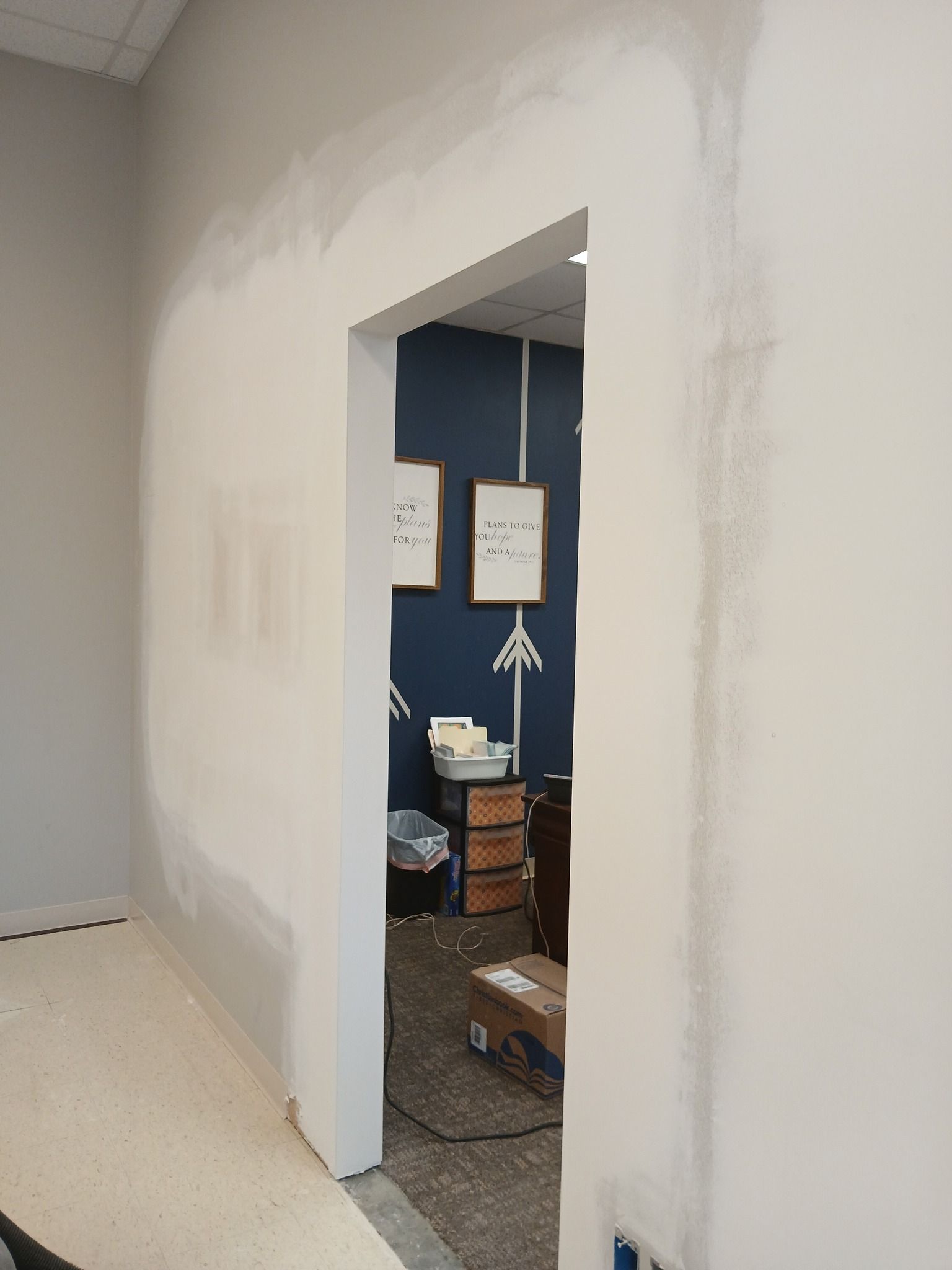White-walled doorway with unfinished drywall.  View into a blue-walled room, two framed papers on wall.