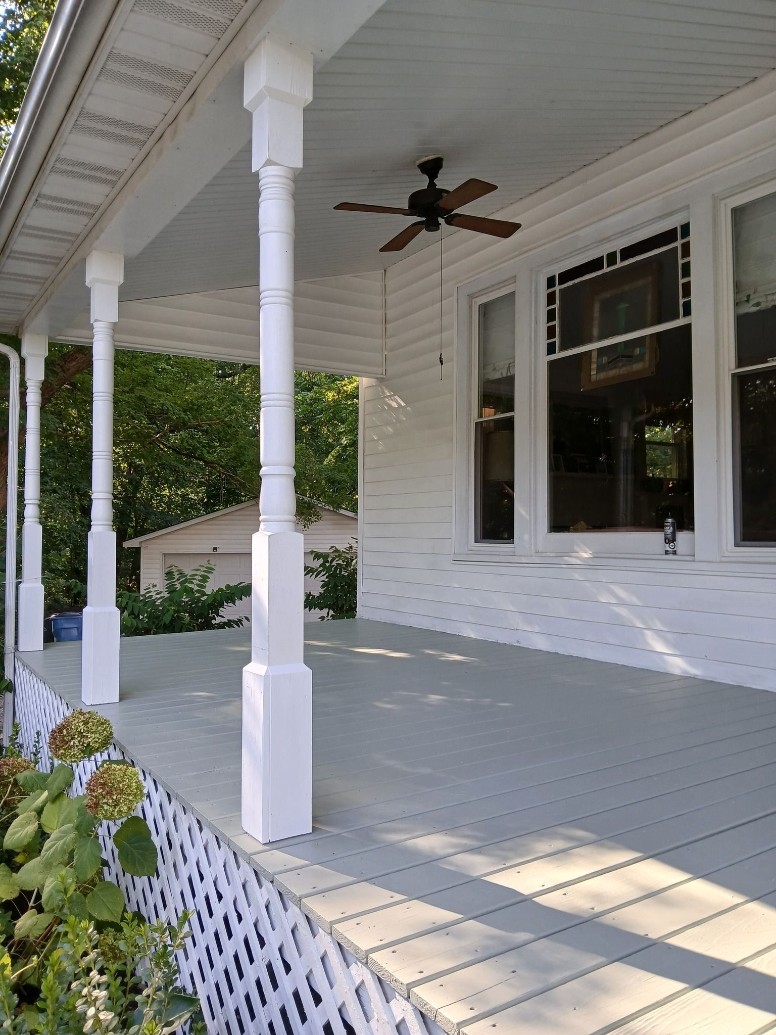White porch with ornate columns, ceiling fan, windows, and light gray floorboards.