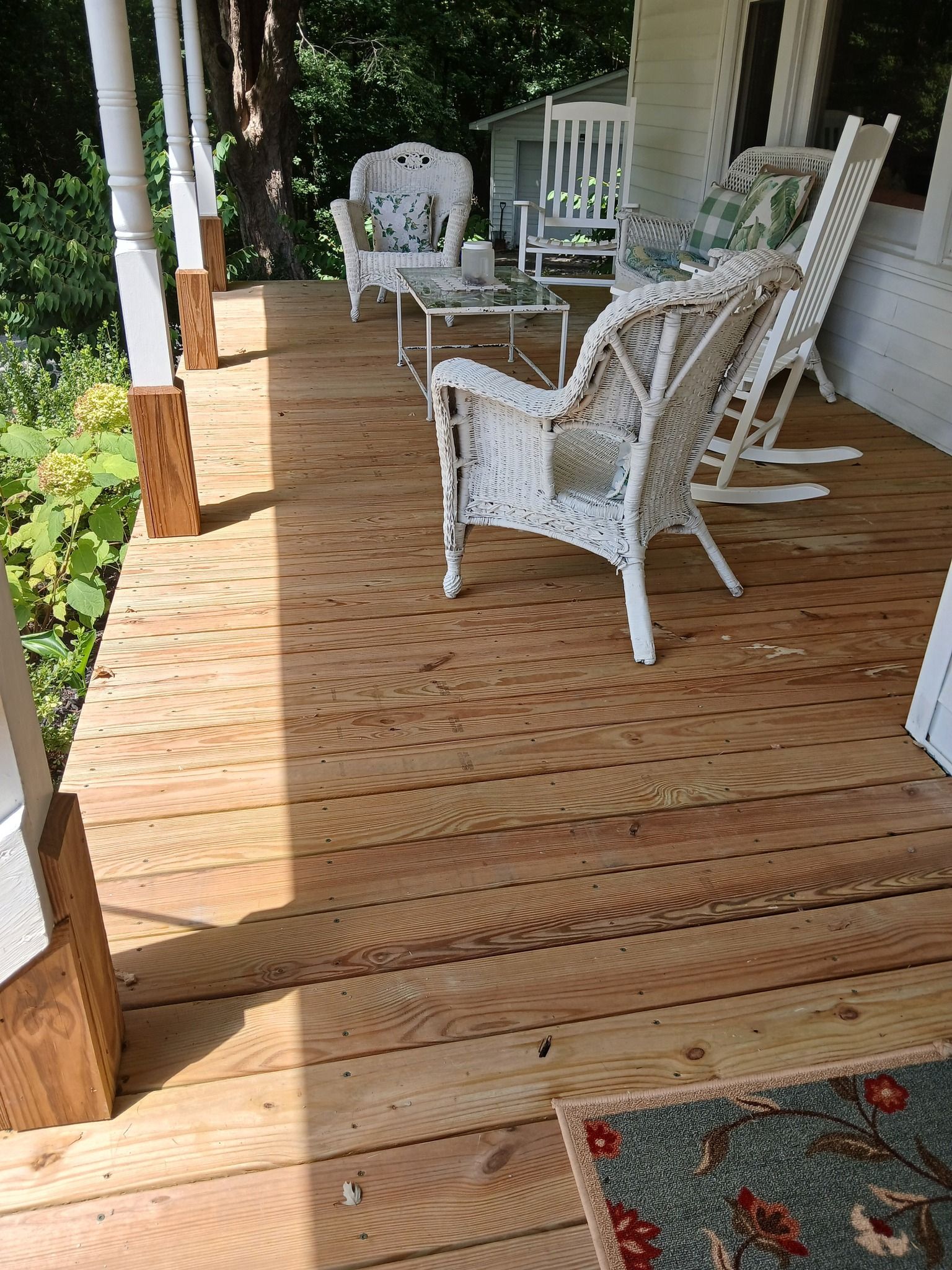 Wooden porch with white wicker furniture; chairs, a table, and a welcome mat.