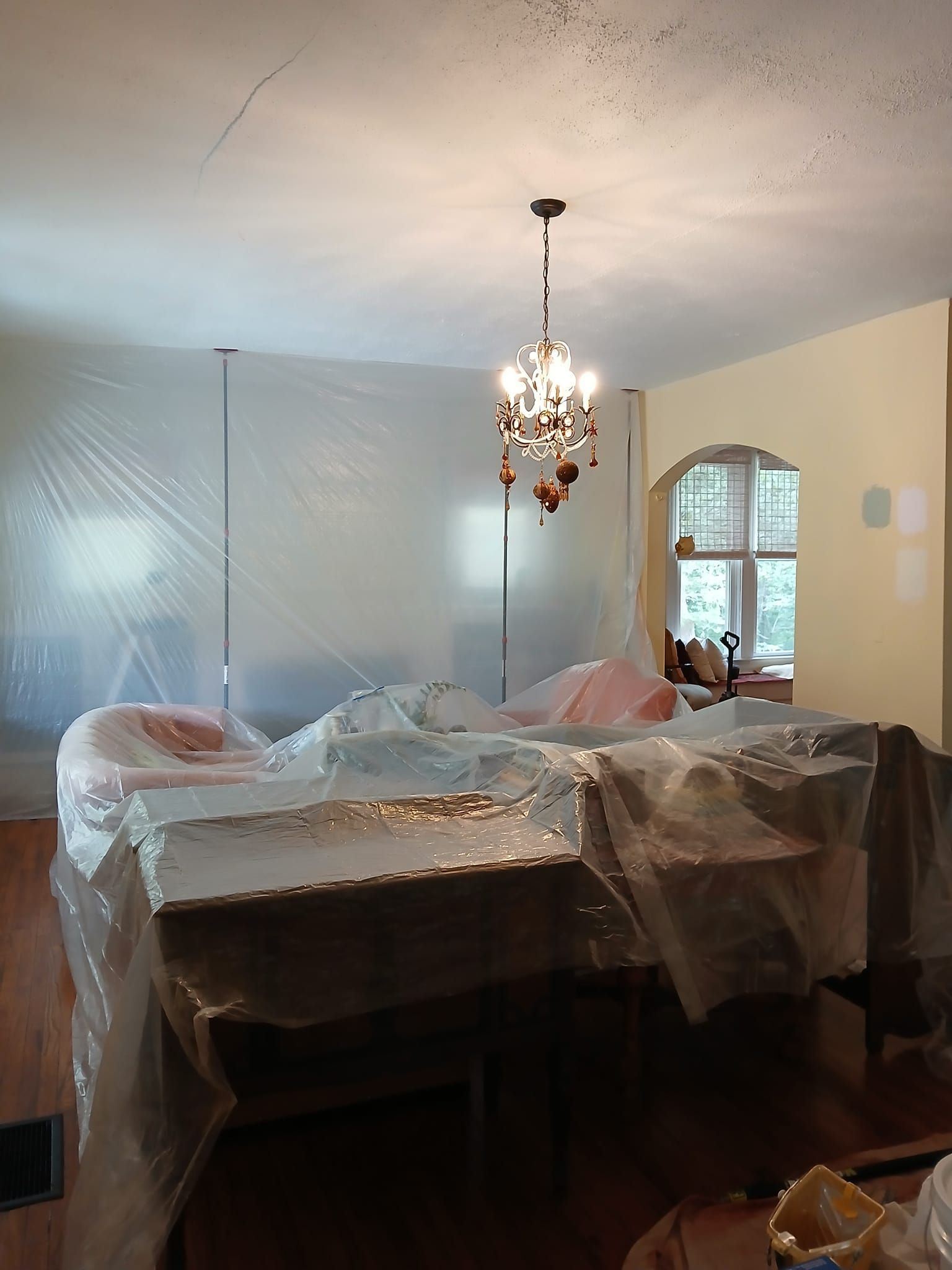 Room being renovated, furniture and walls covered in plastic, chandelier hanging.