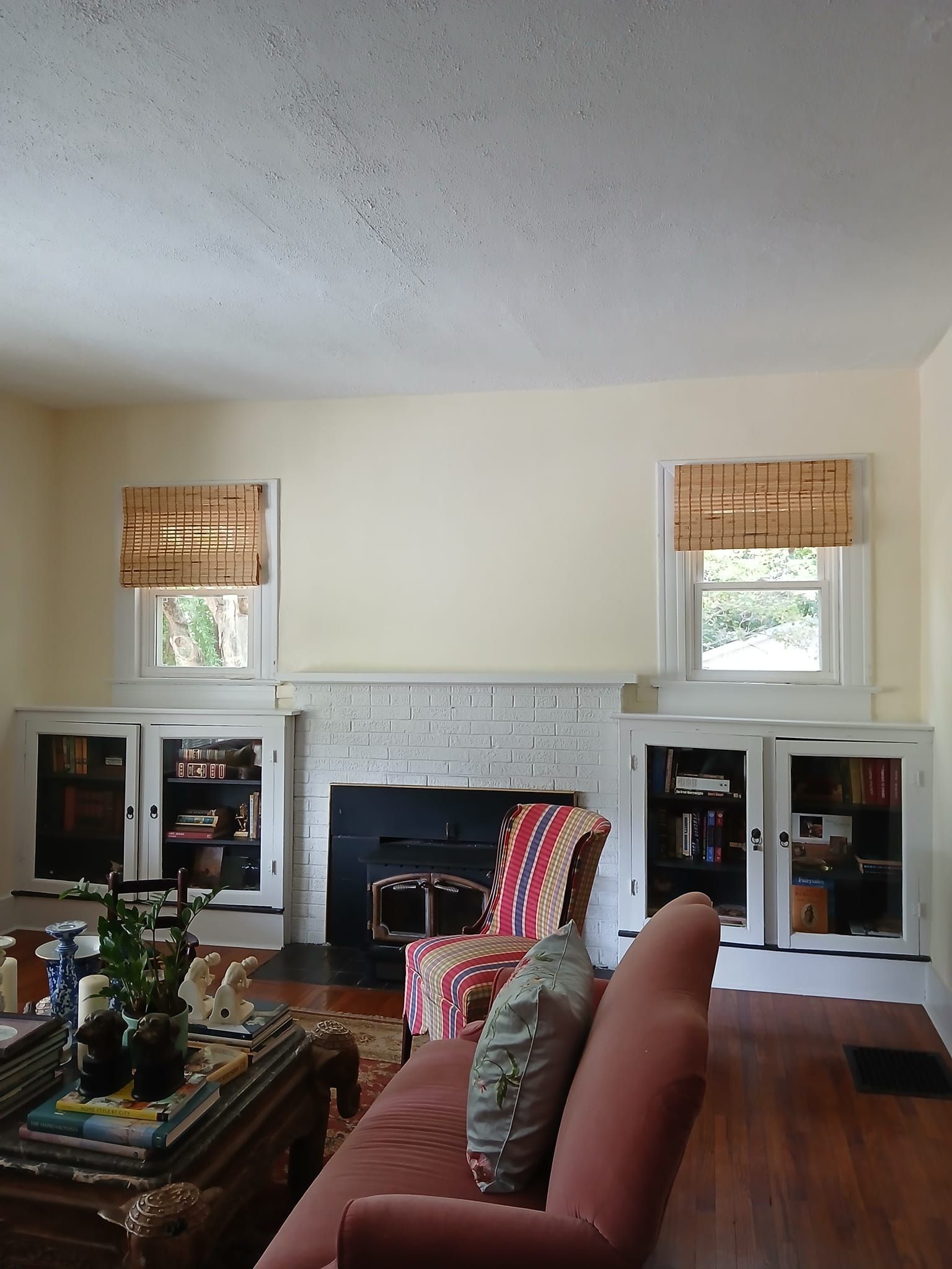 Living room with fireplace, built-in bookshelves, pink sofa, patterned chair, and two windows with woven shades.