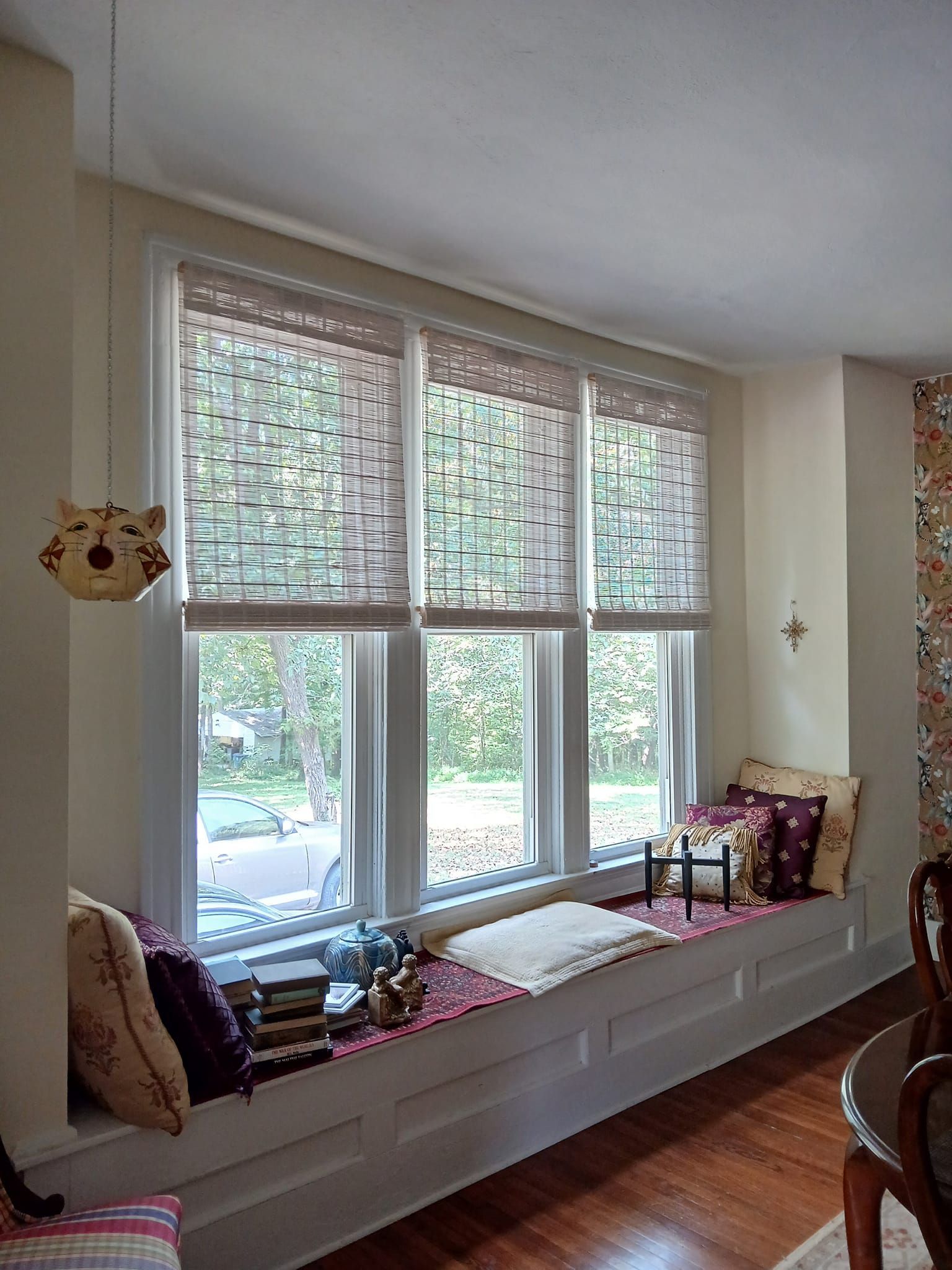 Window seat with three windows, woven shades, pillows, books, and a birdhouse.