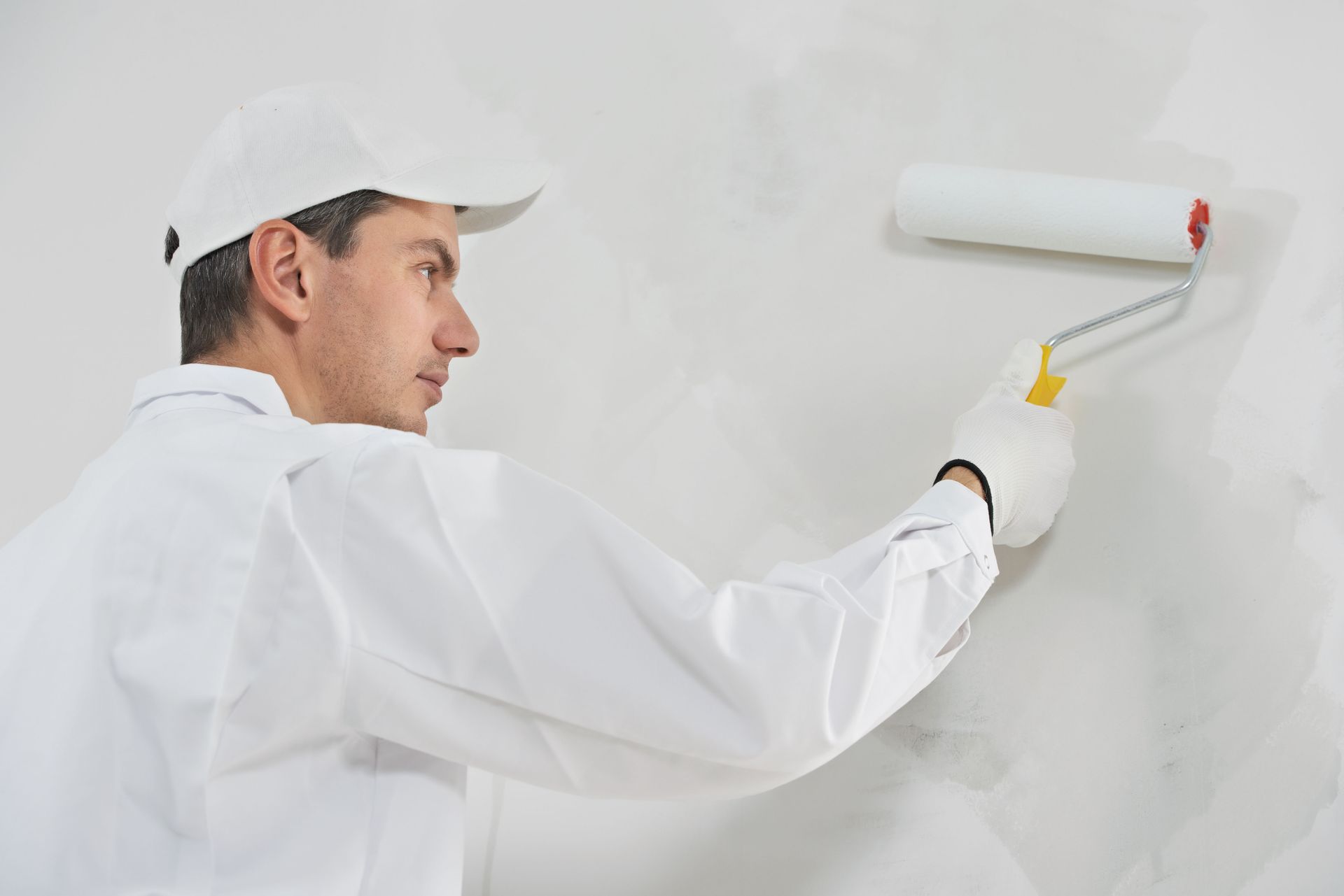 A person in a white uniform and cap painting a white wall with a paint roller.