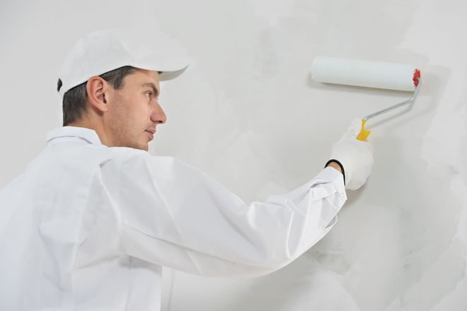 A person in a white uniform and cap painting a white wall with a paint roller.