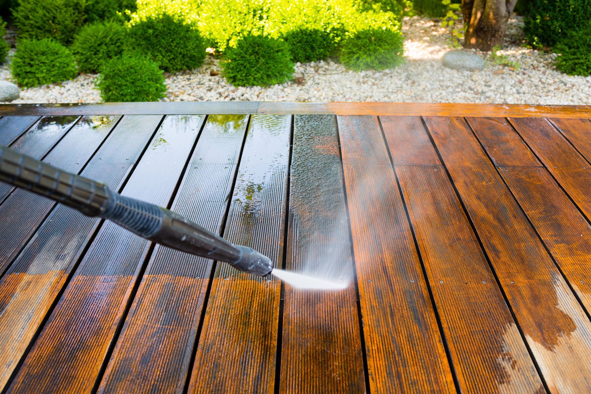 A pressure washer cleans a dirty wooden deck, revealing a contrast between the dark, stained wood and the bright, clean area.