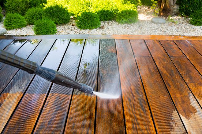 A pressure washer cleans a dirty wooden deck, revealing a contrast between the dark, stained wood and the bright, clean area.