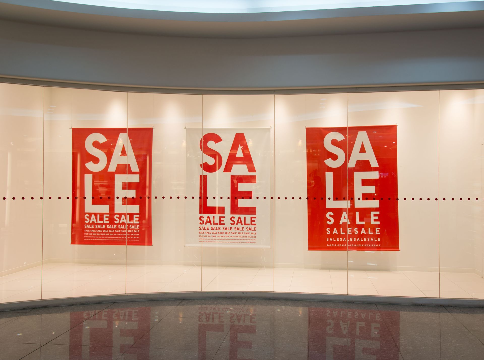 Three red “SALE” posters displayed on a curved mall wall above a reflective floor.