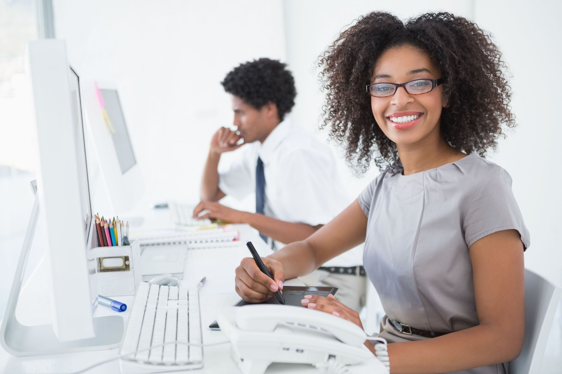 Woman smiling at a desk in a bright office, using a tablet beside a computer and telephone