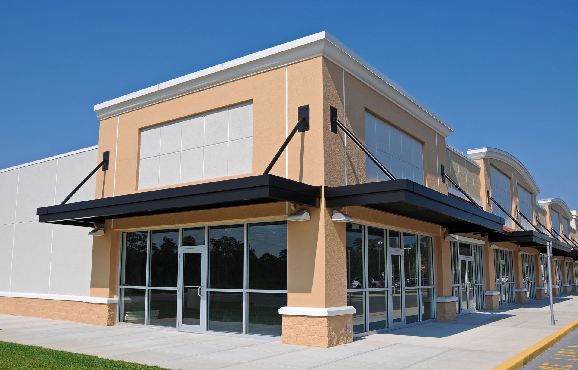 Empty storefronts in a strip mall with tan walls, black awnings, and glass windows on a sunny day