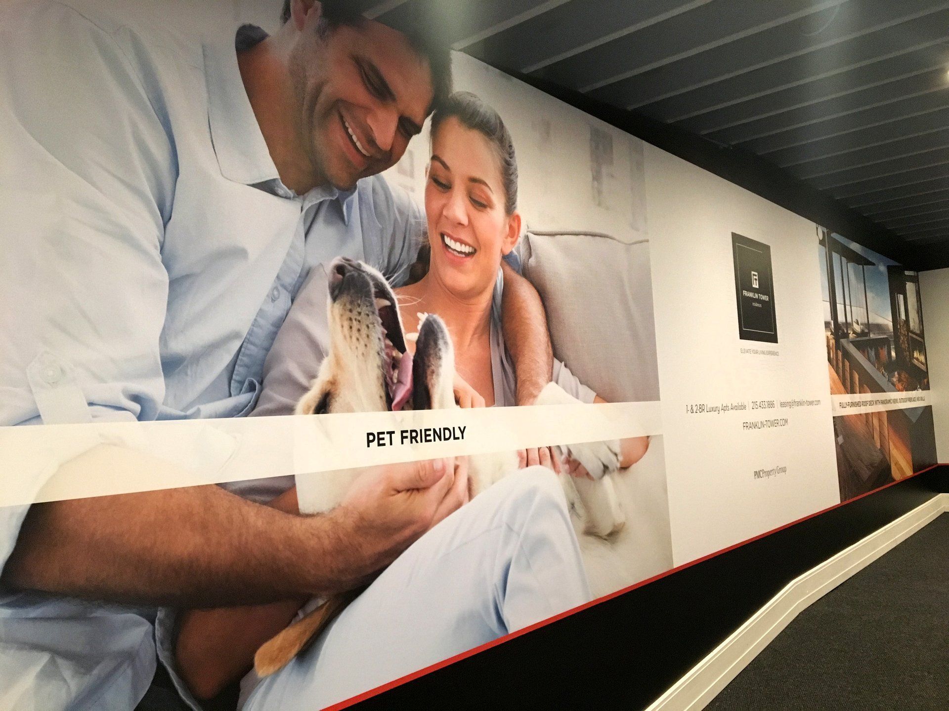 Couple smiling on a fitness-themed wall mural with a small dog, in a bright indoor hallway