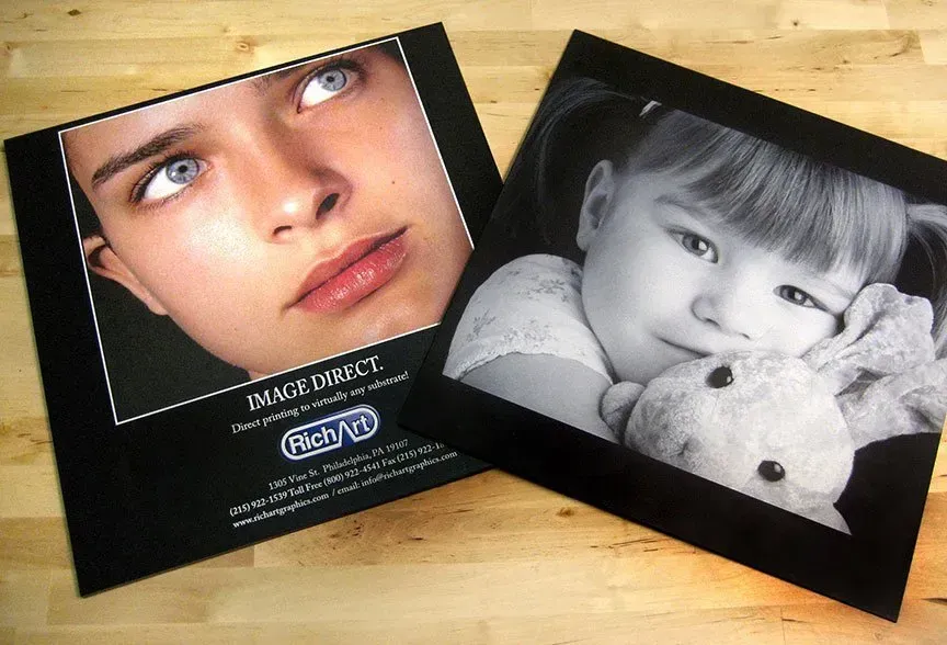 Two black-bordered photo prints on a wooden table, one color portrait and one black-and-white child portrait with a teddy bear.