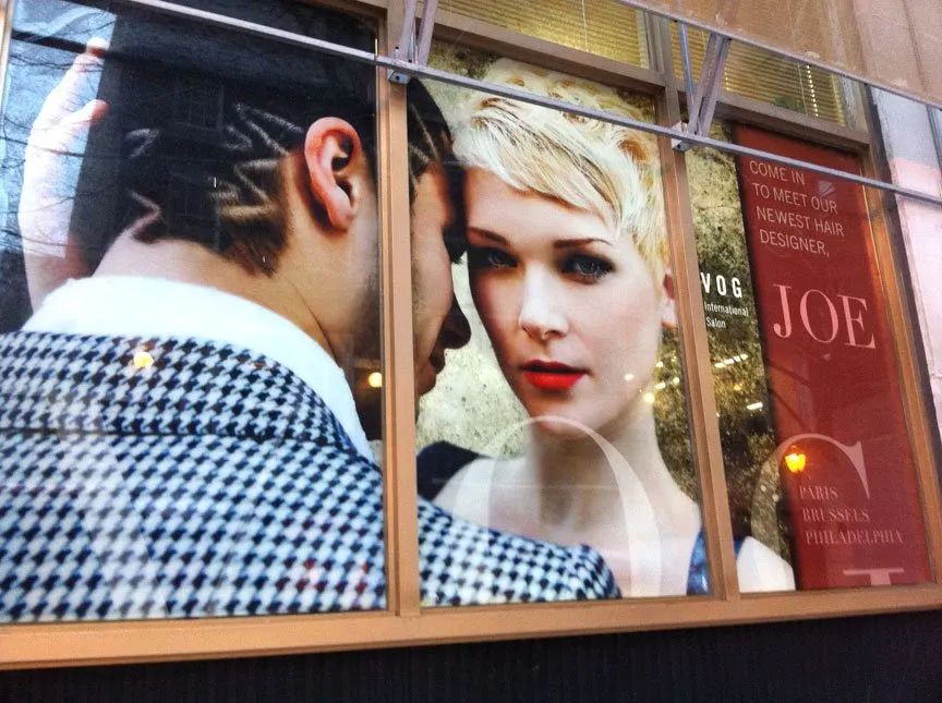 Storefront poster of a man facing a blonde woman with red lips, framed by window panes and the word “JOE” visible