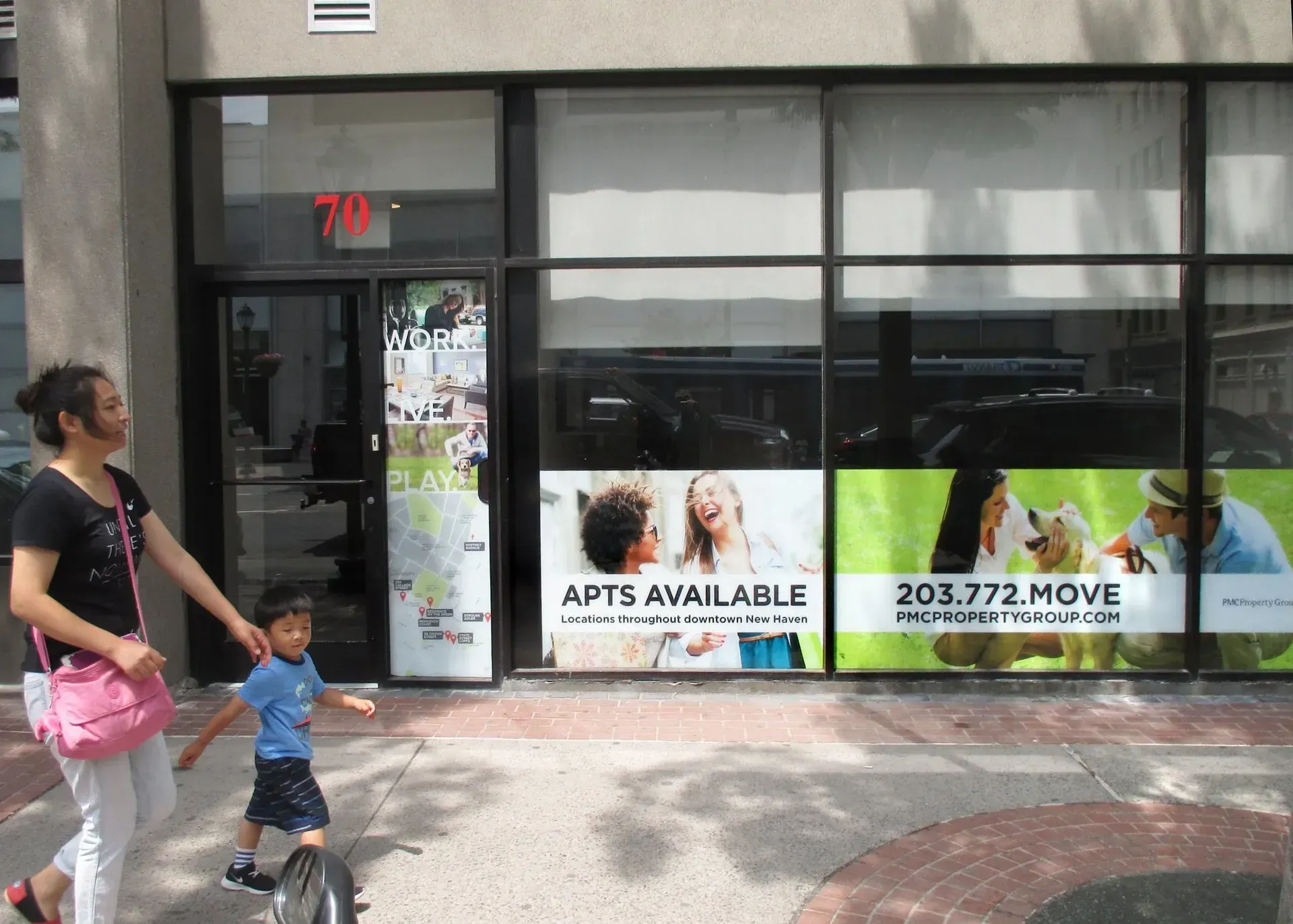 Woman and child walking past a storefront with “APTS AVAILABLE” and “203.772.MOVE” signs.