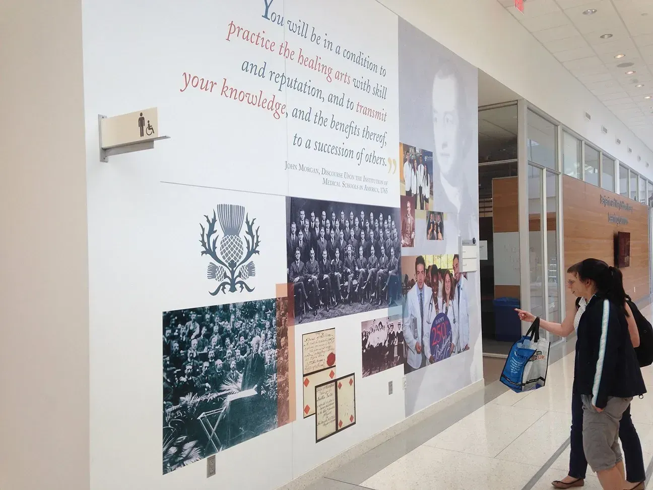 Hallway wall display with photos and text; a person walks past holding a bag.