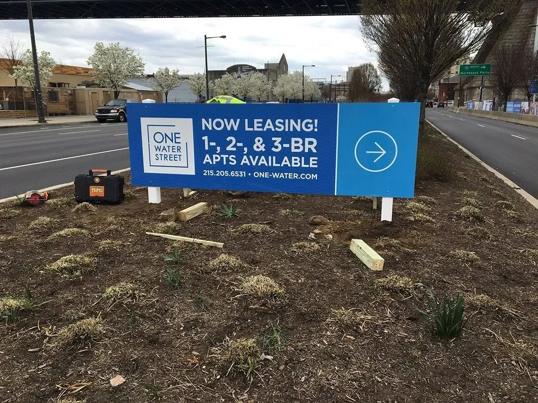 Blue “Now Leasing” sign for 1-, 2- and 3-bedroom apartments in a roadside planter bed.