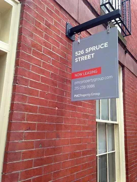 Gray hanging sign on a red brick building reads “526 Spruce Street” and “Now Leasing.”