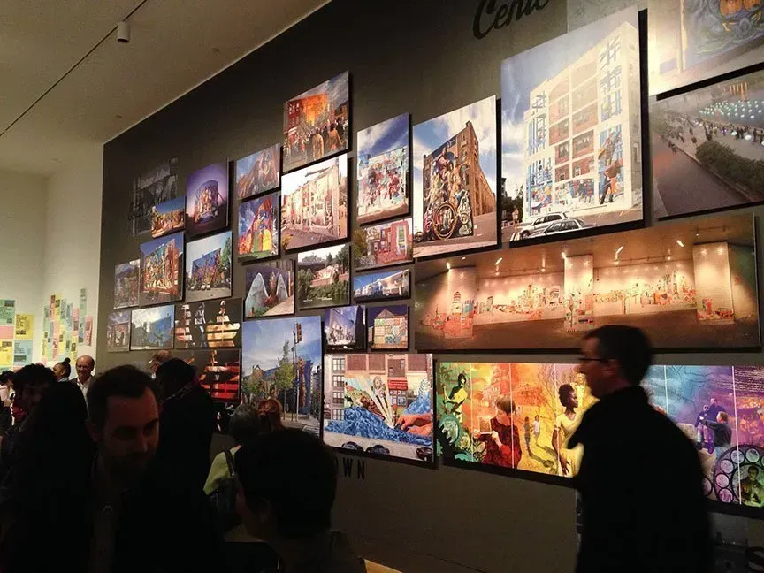 Large wall of colorful art prints in a gallery, with visitors standing and walking by.