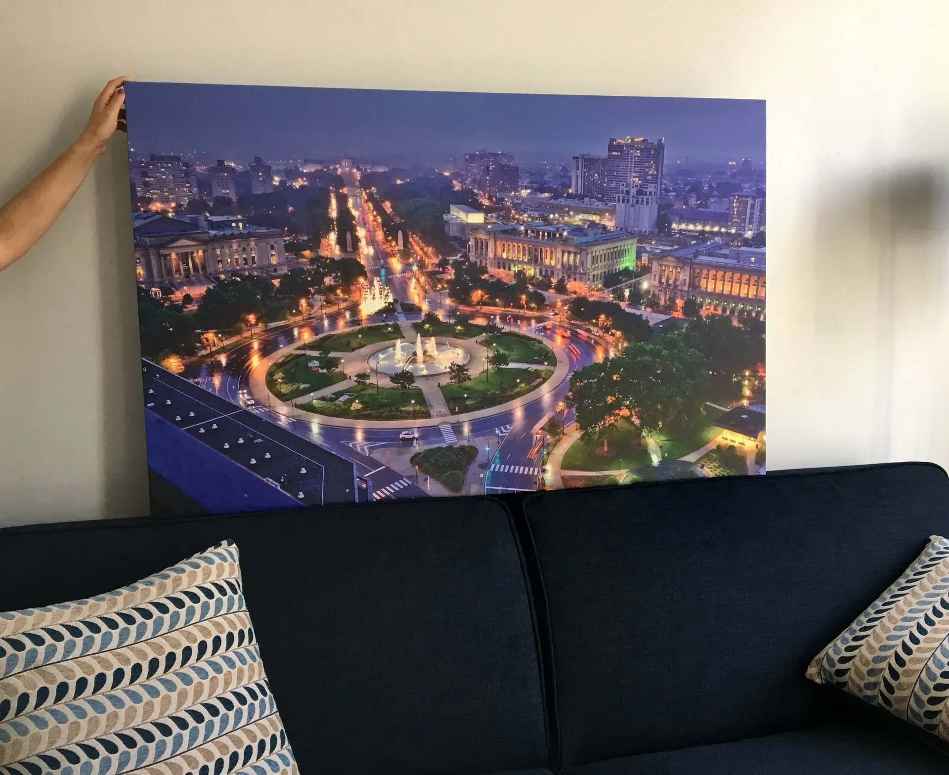 Framed night cityscape above a dark sofa with patterned cushions, lit by warm streetlights and buildings