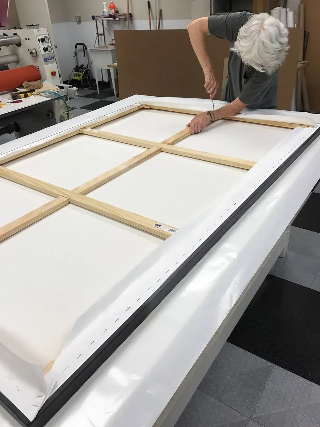 Person stretching fabric over a large white canvas frame in a workshop