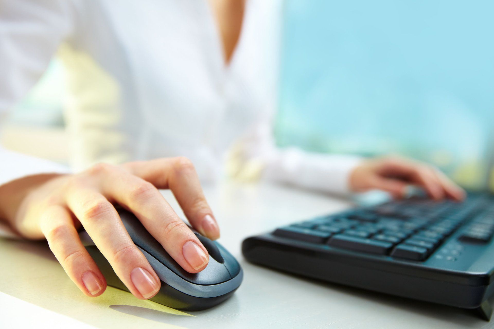 Hand on a mouse beside a keyboard on a desk, with a blurred person in the background
