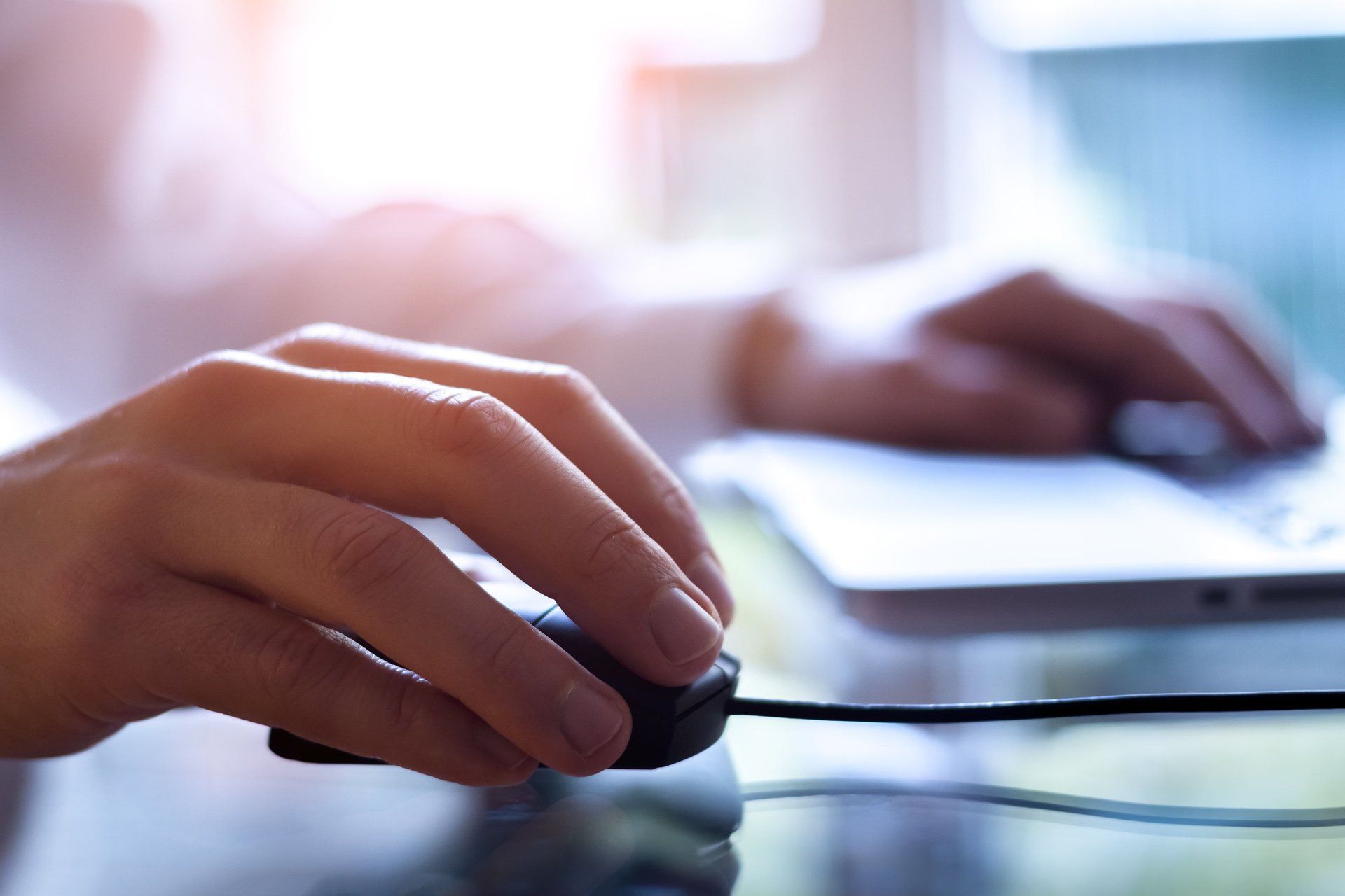 Hand on a mouse beside a laptop, working at a desk in soft light
