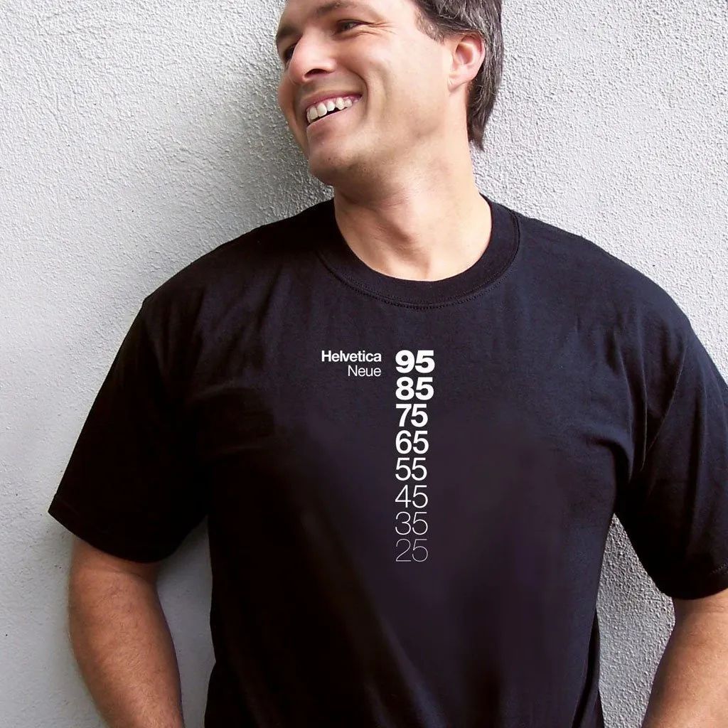 Smiling man in black T-shirt with vertical white numbers against a textured gray wall