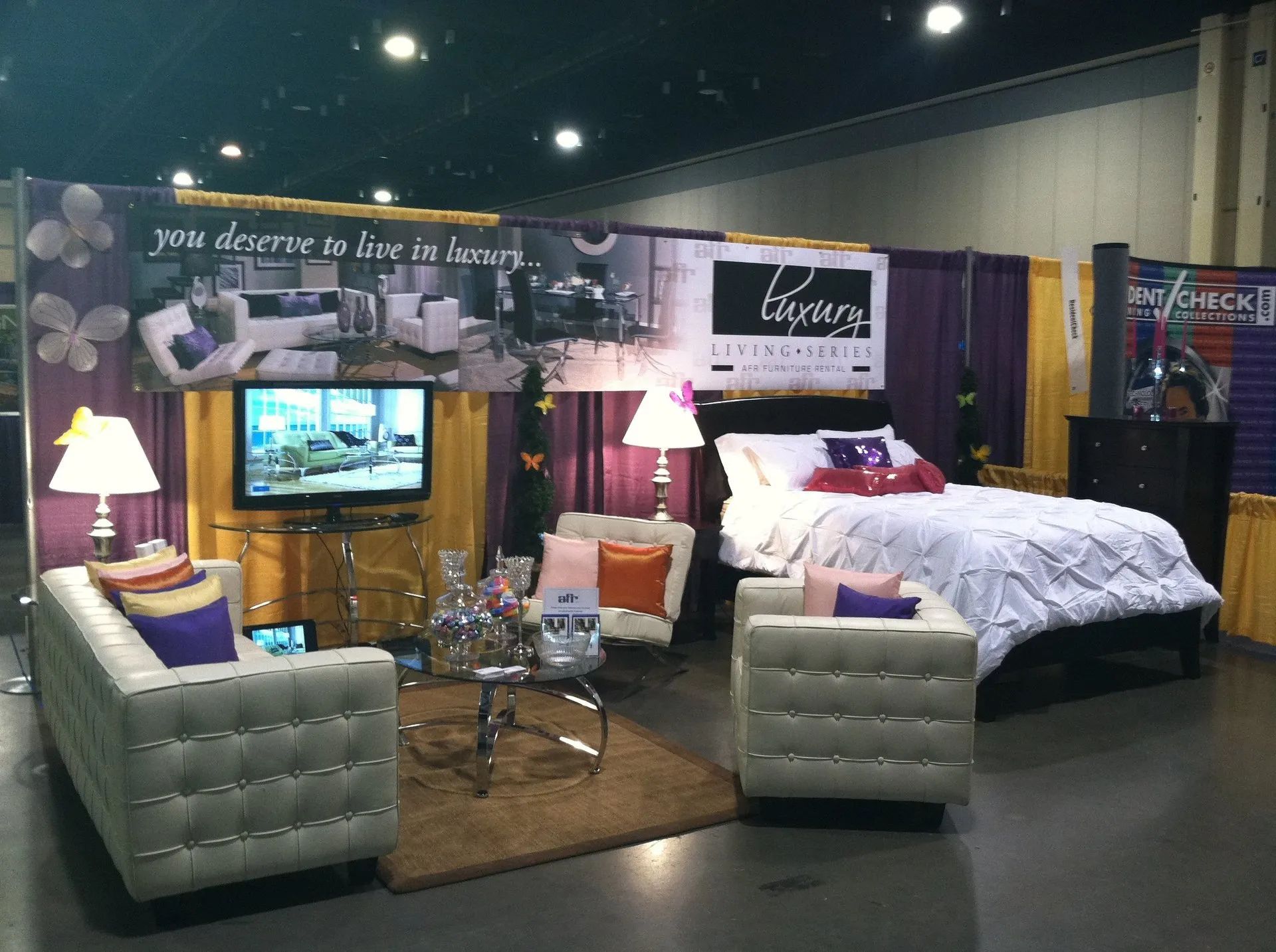 Trade show bedroom display with a white bed, tufted chairs, TV, and purple lamps on a carpeted booth.