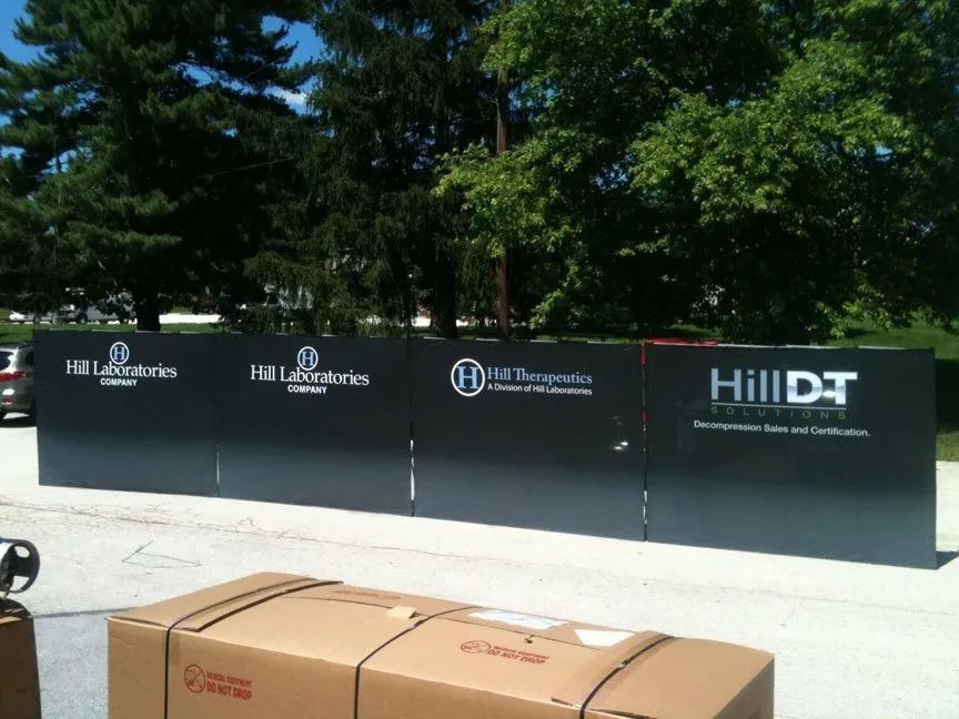 Black fence with Hill DT logos in front of trees, with cardboard boxes on the ground in the foreground
