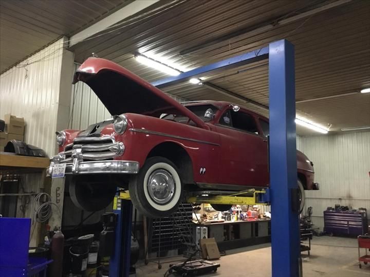 A red vintage car with its hood open, raised on a blue automotive lift inside a repair garage.