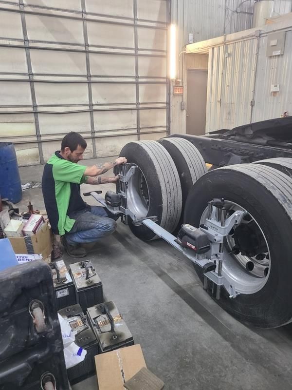 A technician in a green and black uniform attaches diagnostic alignment sensors to the rear wheels of a semi-truck.