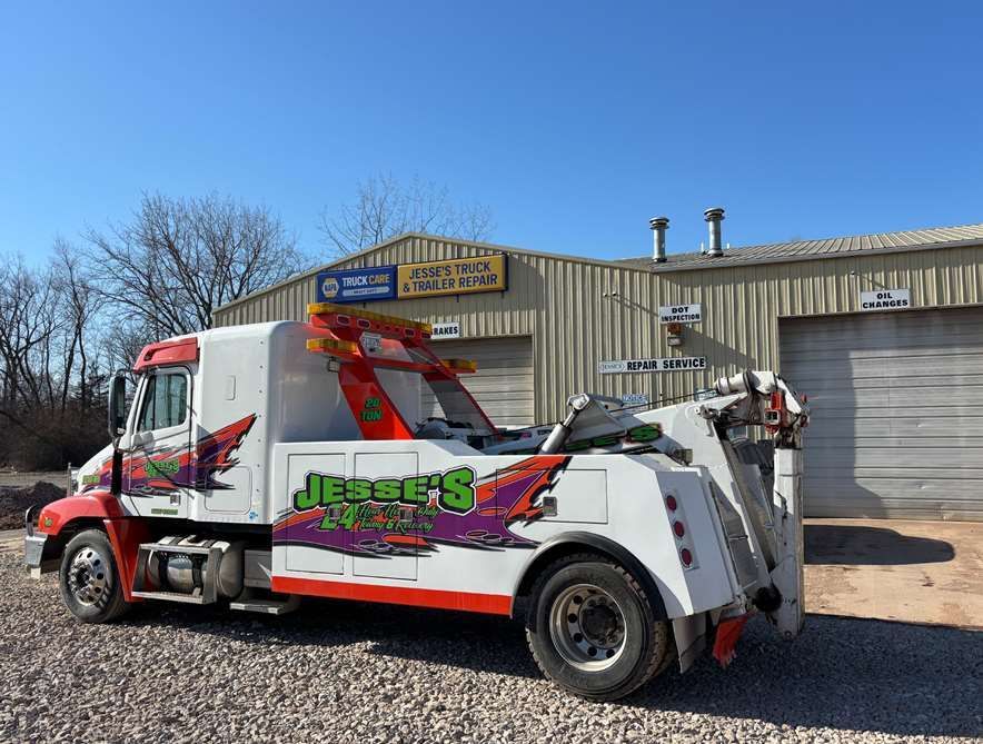 A white Jesse's 24 Hour Towing truck parked on a gravel lot in front of a metal warehouse building under a blue sky.