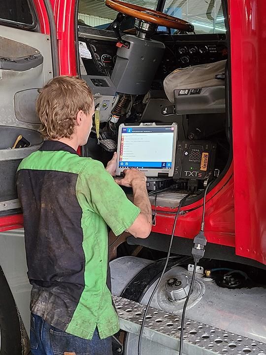 A mechanic wearing a green and black uniform uses a diagnostic laptop inside the open cab of a red truck.
