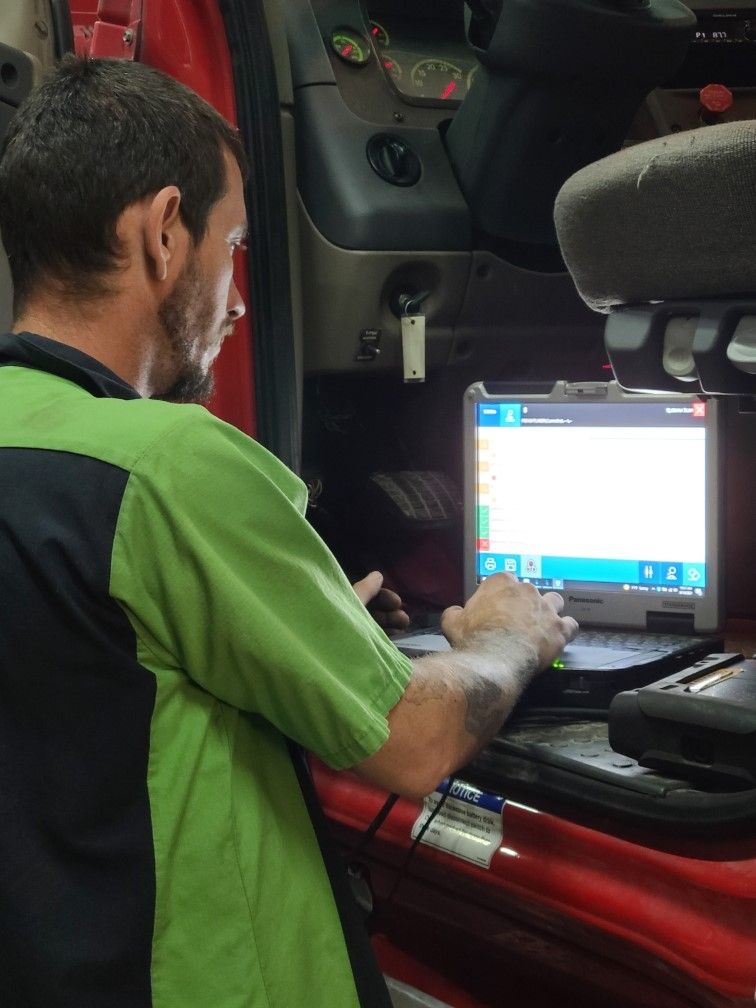 A mechanic in a lime green shirt sits inside a vehicle cab, operating a diagnostic laptop connected to the truck.