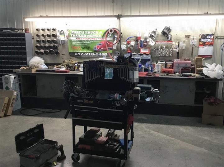A cluttered mechanic's workshop featuring a central rolling tool cart, a workbench with tools, and overhead lighting.