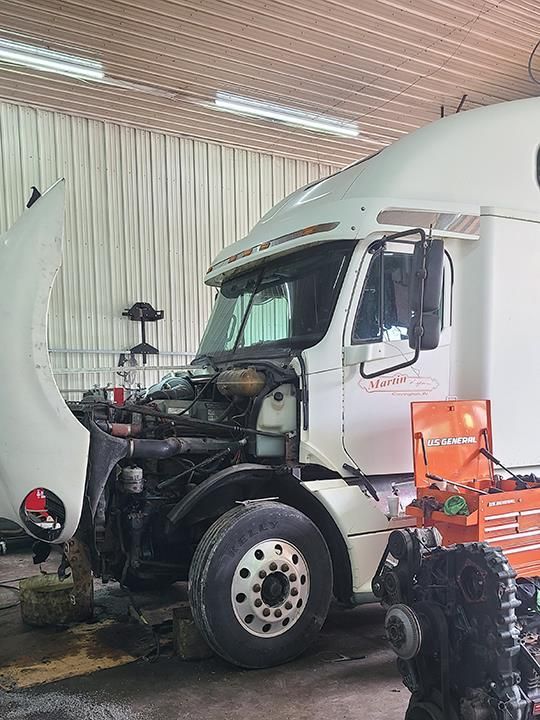 A white semi-truck in a garage with its hood open for maintenance, positioned next to a mechanic's orange tool chest.
