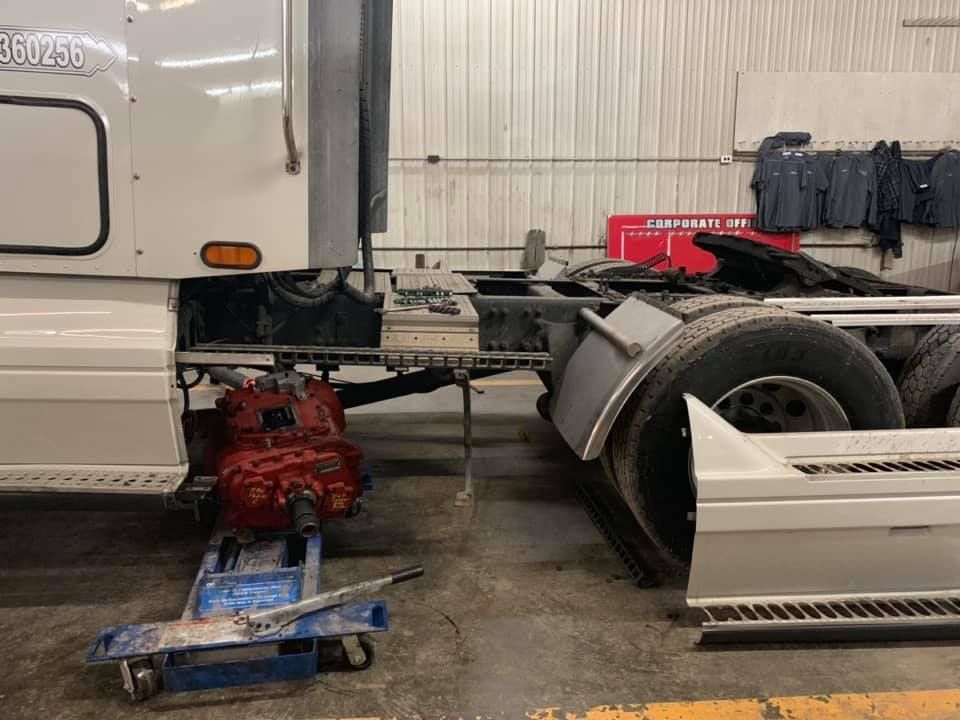 A white semi-truck in a garage with a red transmission removed and supported on a blue jack stand.