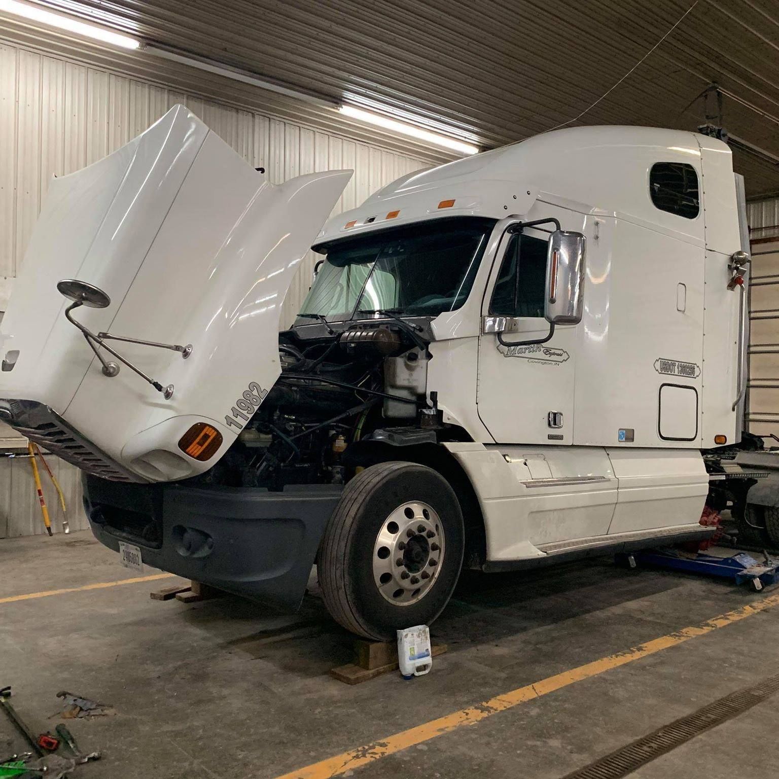 A white semi-truck with its hood open in a garage, resting on wooden blocks.