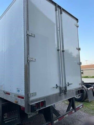 Rear view of a white box truck trailer with double doors and metal locking hardware, parked on a gravel lot.