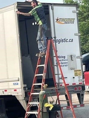 A worker stands on the top step of a ladder to access the upper frame of an open semi-trailer, with a gas cylinder below.