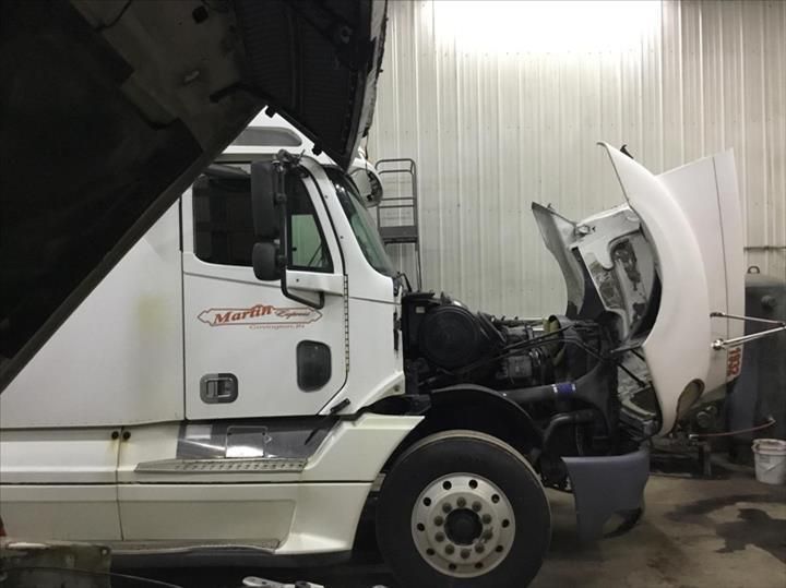 A white semi-truck parked in a garage with its hood open for engine maintenance.