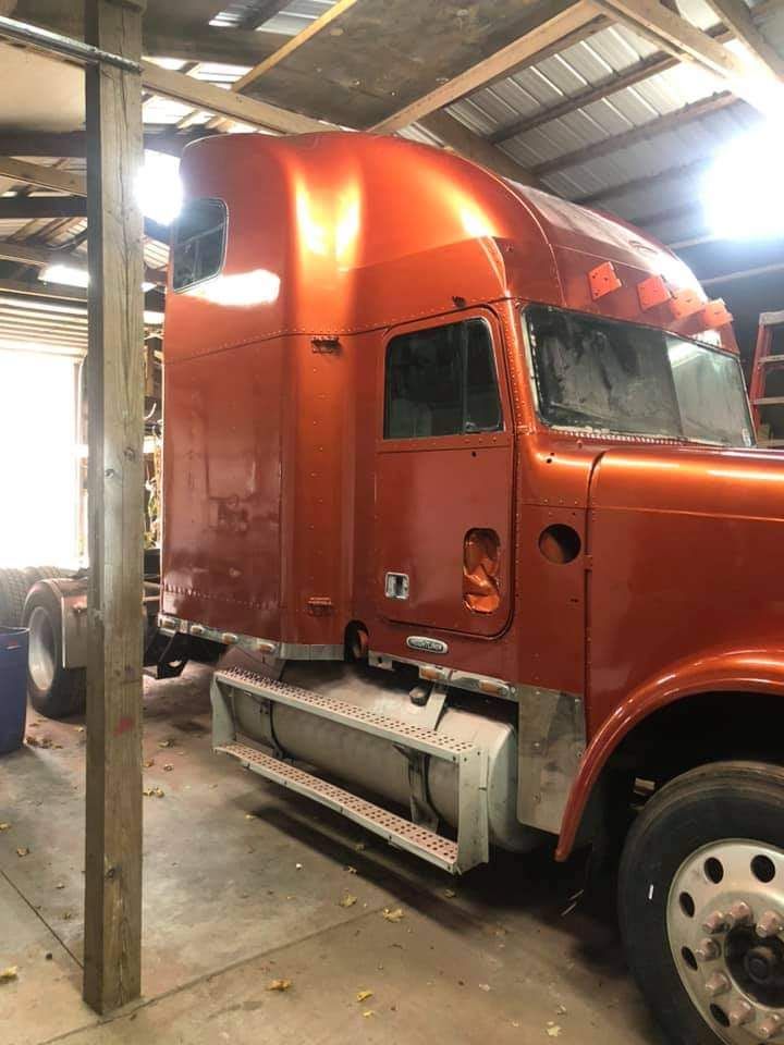 A freshly painted metallic orange semi-truck cab sits inside a dimly lit workshop.