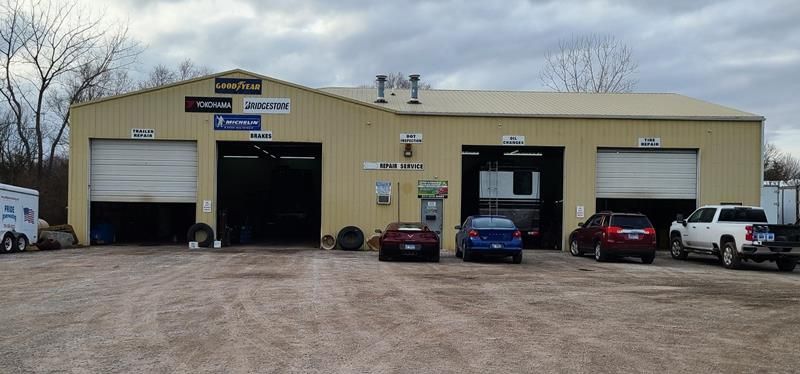 A tan auto repair shop with multiple garage bays, tire brand signs, and several parked cars in a gravel lot.