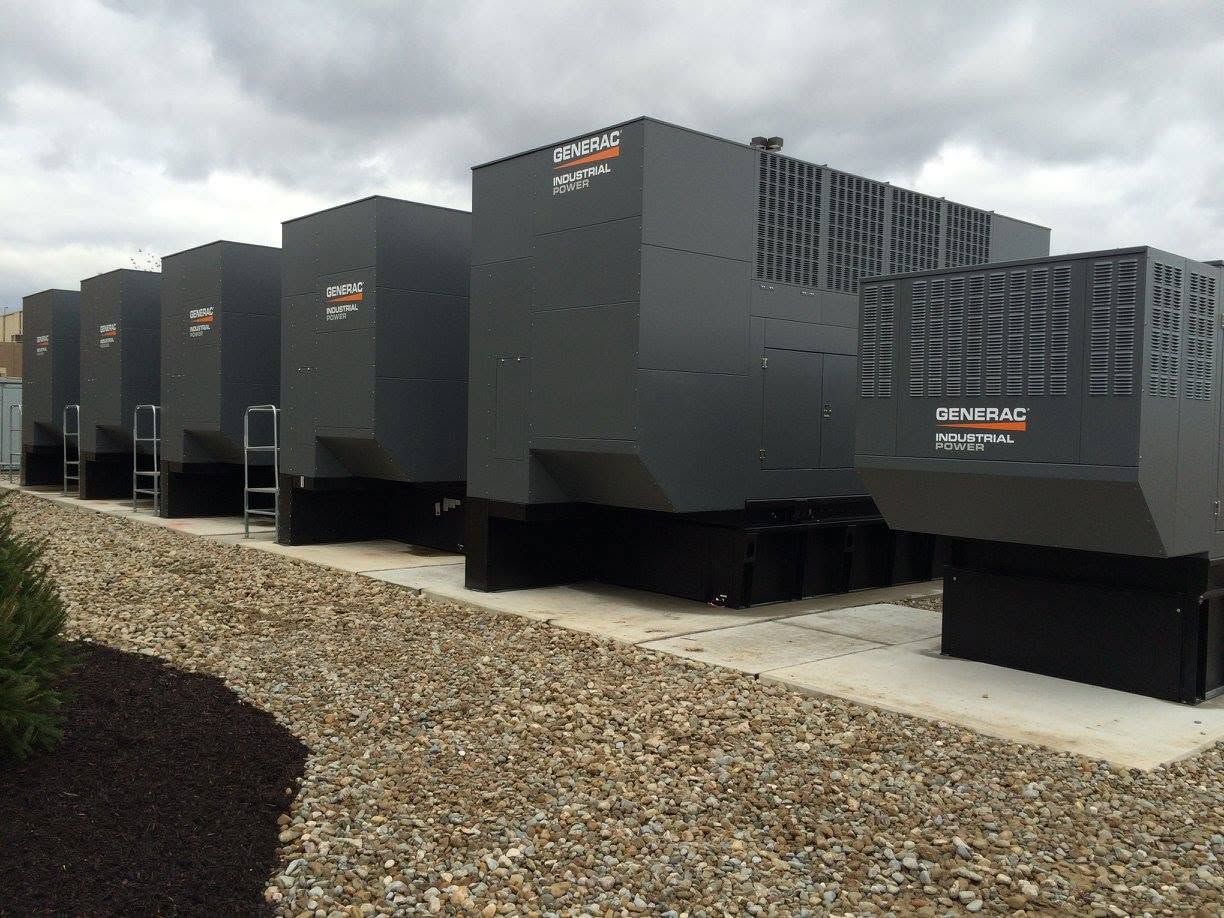 Row of large dark industrial cooling units on a gravel lot under a cloudy sky