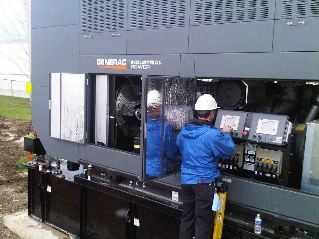 Worker in hard hat inspecting controls on a large black industrial generator outdoors.