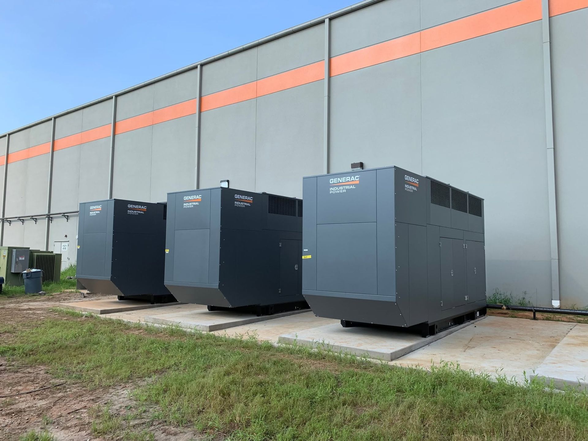 Three large gray industrial generators beside a concrete wall outdoors.
