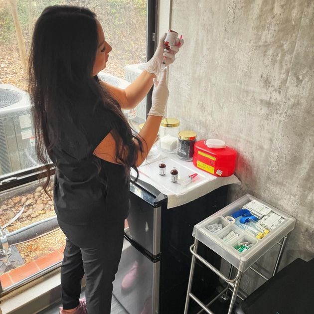 A medical professional in black scrubs and gloves draws medication from a vial in a clinical office setting.